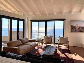 Amplo quarto com uma grande porta de vidro deslizante que dá para uma varanda com vista para o oceano. Há um sofá de couro, duas poltronas e uma mesa de centro de madeira.