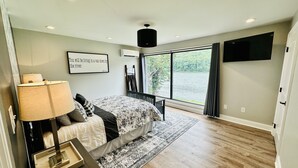 Un dormitorio con cama, una mesita de noche con lámpara, un ventanal con vista a un río y un televisor montado.