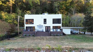 Una casa moderna de dos pisos con un balcón y una zona cubierta, ubicada a orillas de un río con ribera rocosa.