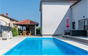 Ein Pool mit überdachtem Pavillonbereich, ein modernes Gebäude und Sitzmöglichkeiten im Freien.