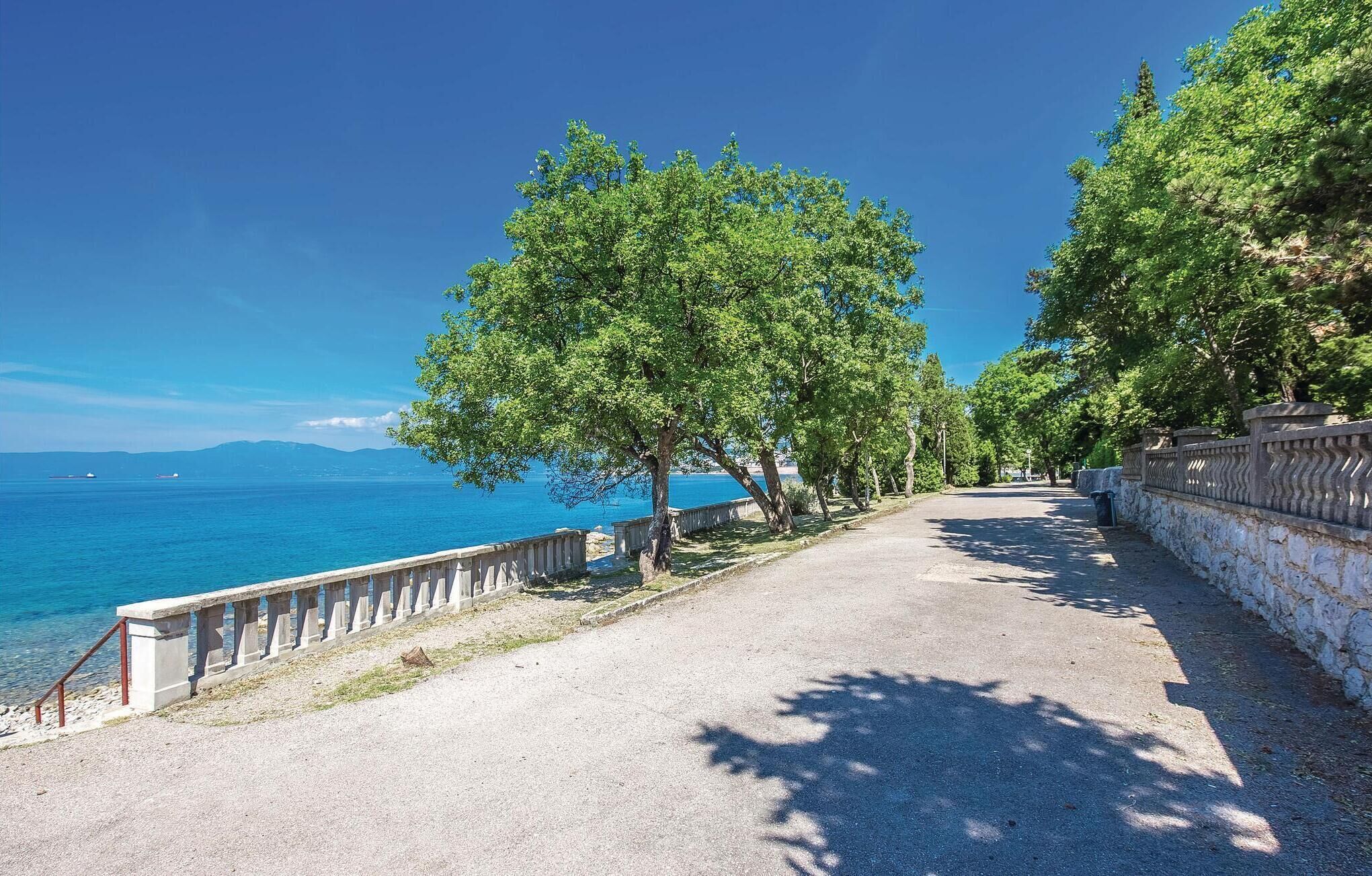 A paved pathway with a stone wall on one side and a railing on the other, leading towards a body of water with trees lining the path.