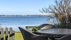 Un patio con mobiliario de mimbre que da a un lago, una cerca de madera y una vista distante de un pueblo.