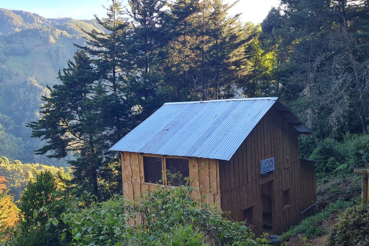 A wooden cabin with a metal roof, surrounded by dense forest and mountains.