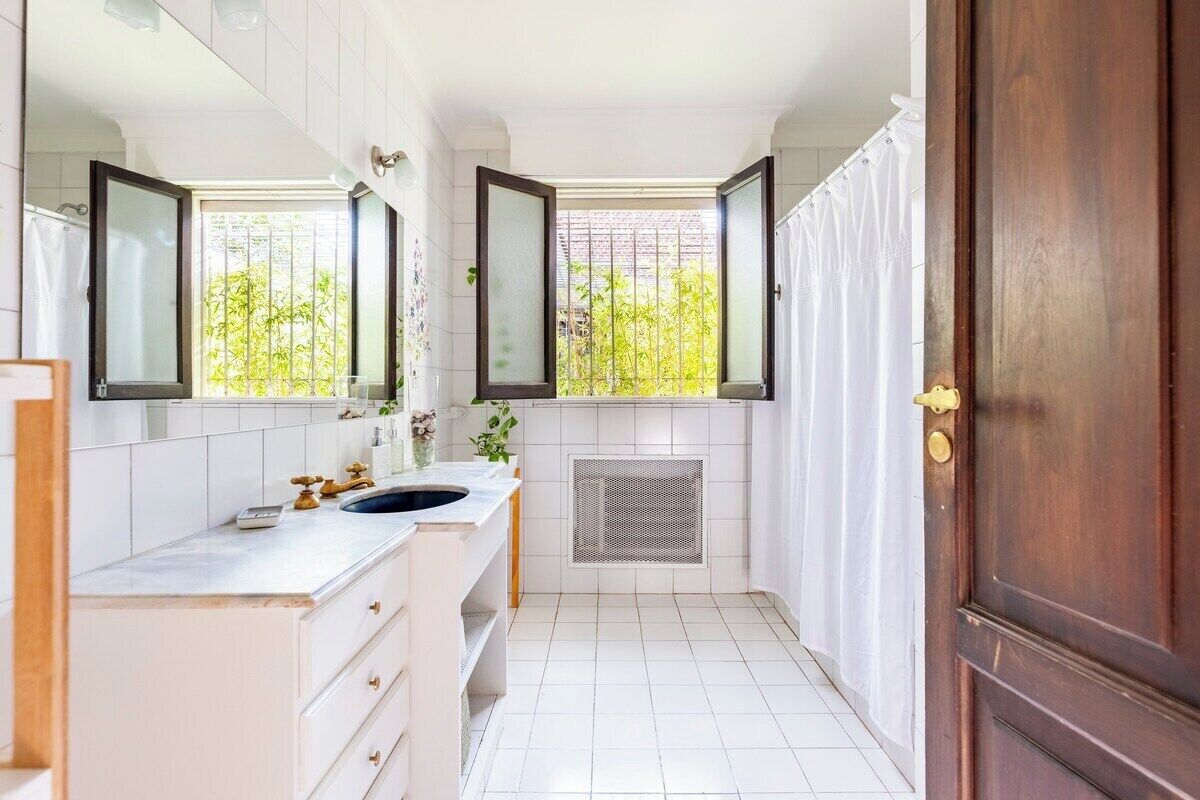 A narrow bathroom with white tiles, a double sink, a mirror, and a window with a view of greenery.