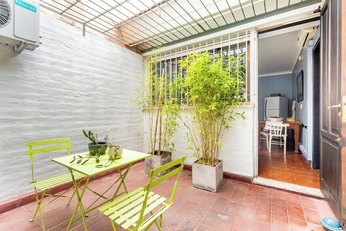 Un pequeño patio amurallado con una mesa, sillas, plantas en macetas y una ventana con barrotes.
