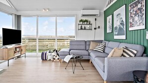 Een moderne woonkamer met een grijze bank, een flatscreen-tv en uitzicht op het landschap door grote ramen.