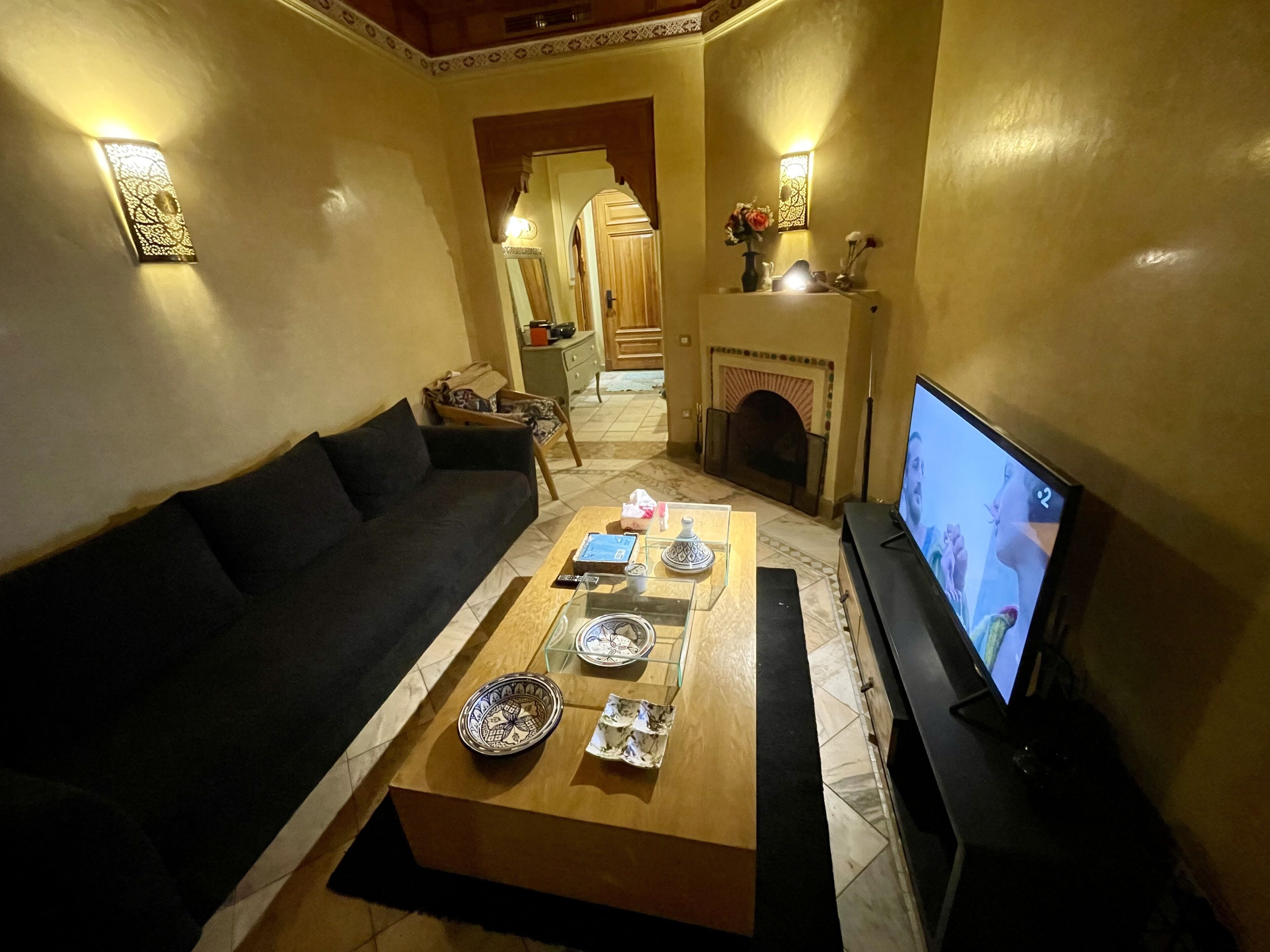 A living room with a sofa, coffee table, and TV. There is a fireplace and a hallway leading to a kitchen.