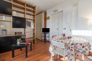 Une pièce avec une bibliothèque en bois, un téléviseur à écran plat, une table à manger recouverte d’une nappe à motifs floraux et un panneau indiquant « ZONE FÊTE ».