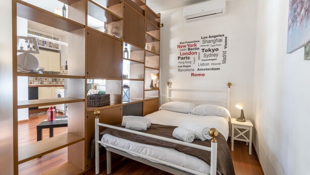 Un dormitorio ordenado con cama, mesitas de noche, armario y una pared con una nube de palabras que muestran nombres de ciudades.