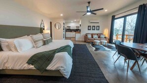 Un dormitorio con cama, un comedor con mesa y sillas, una cocina al fondo y un ventanal grande.
