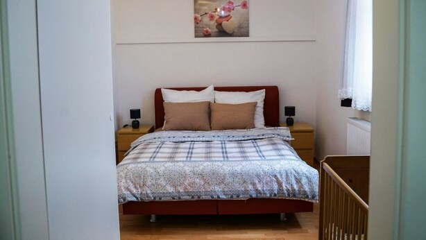 A bedroom with a bed, two bedside tables, a nightstand, a wall-mounted lamp, and a window with curtains.