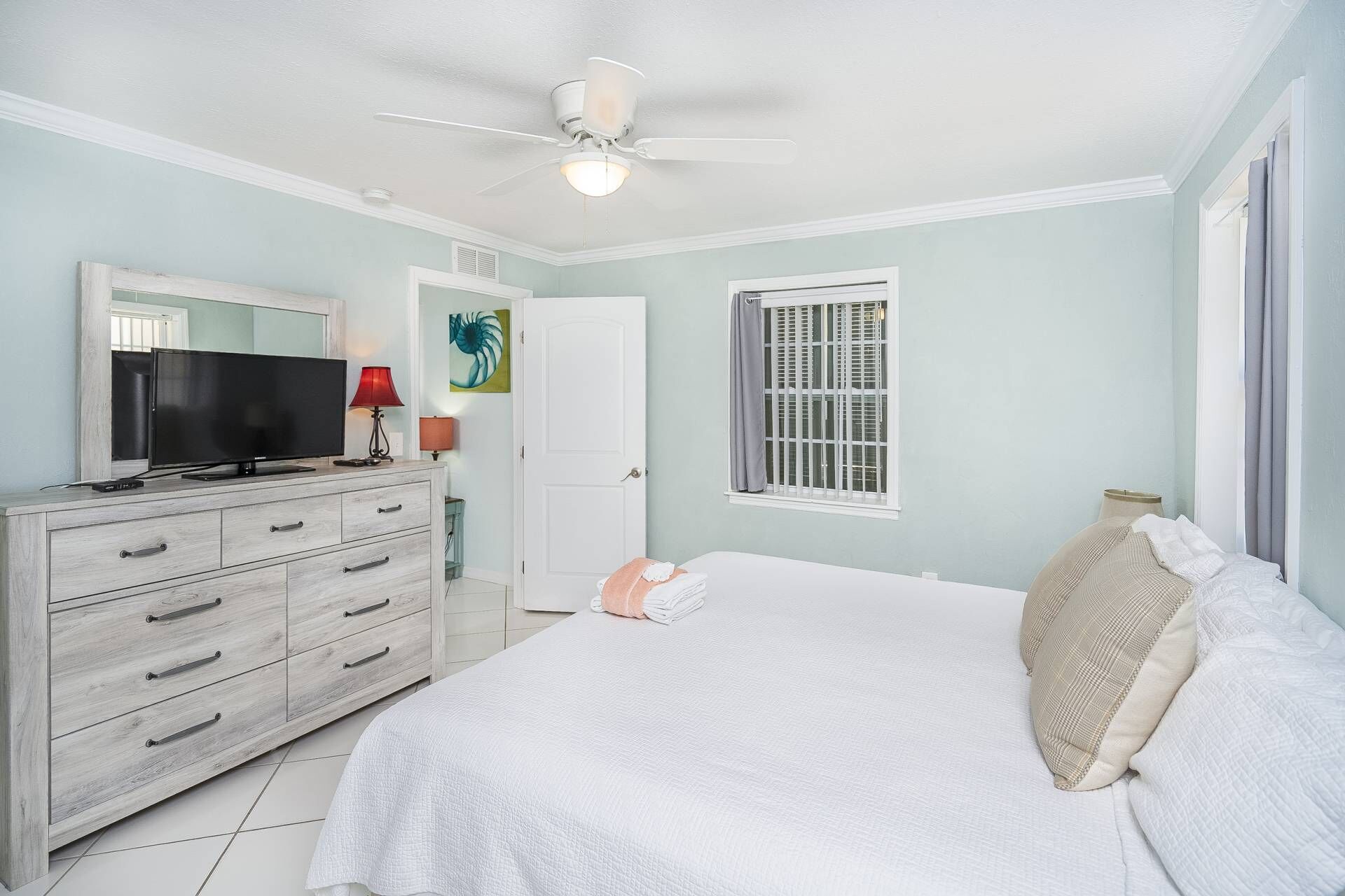 Une chambre à coucher comprenant un lit, un téléviseur, une commode, un ventilateur de plafond et une fenêtre avec des volets.