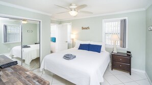 A bedroom with a bed, a nightstand, a ceiling fan, a window with curtains, and a mirror. - Oceans 13 Unit B (St. Augustine)