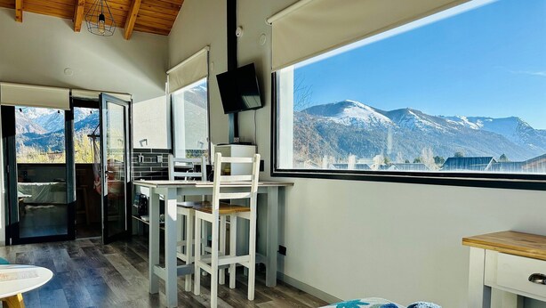 Un dormitorio con cama, escritorio y silla, una ventana con vista a la montaña, y vista a la cocina y el comedor.