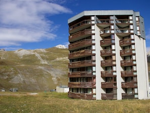 Um edifício de vários andares com varandas, com montanhas ao fundo.