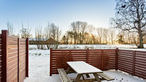 Una mesa de picnic de madera con un banco, rodeada por una valla de madera, ubicada en un área exterior nevada con árboles y cielo despejado.
