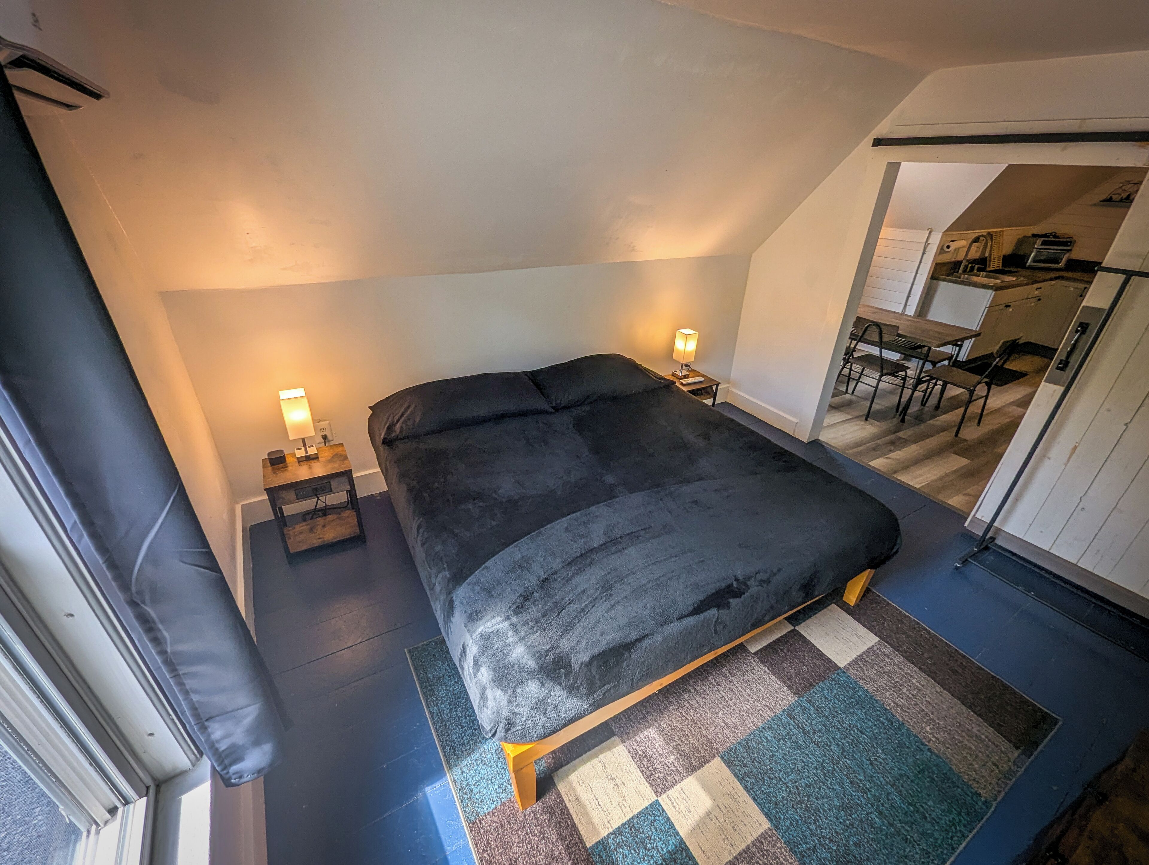 A bedroom with a large bed, two bedside tables with lamps, a small desk, and a kitchenette in the background.