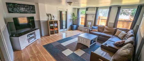 Ein modernes Wohnzimmer mit Flachbildfernseher, Ecksofa, Couchtisch und großem Fenster mit Blick ins Freie.