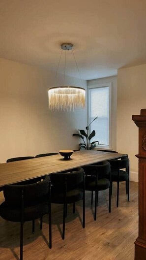 Un espace repas comprenant une longue table en bois, des chaises noires, un luminaire suspendu et une fenêtre avec des stores.