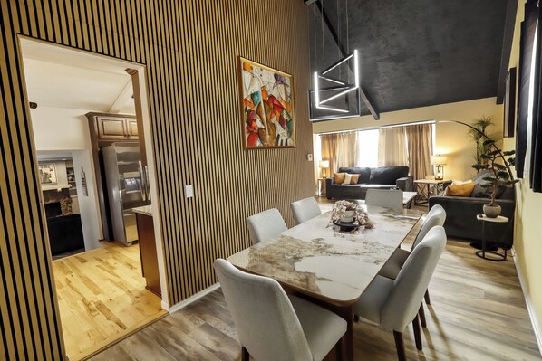Un comedor moderno con una mesa grande y sillas, una obra de arte contemporánea en la pared y una cocina al fondo.