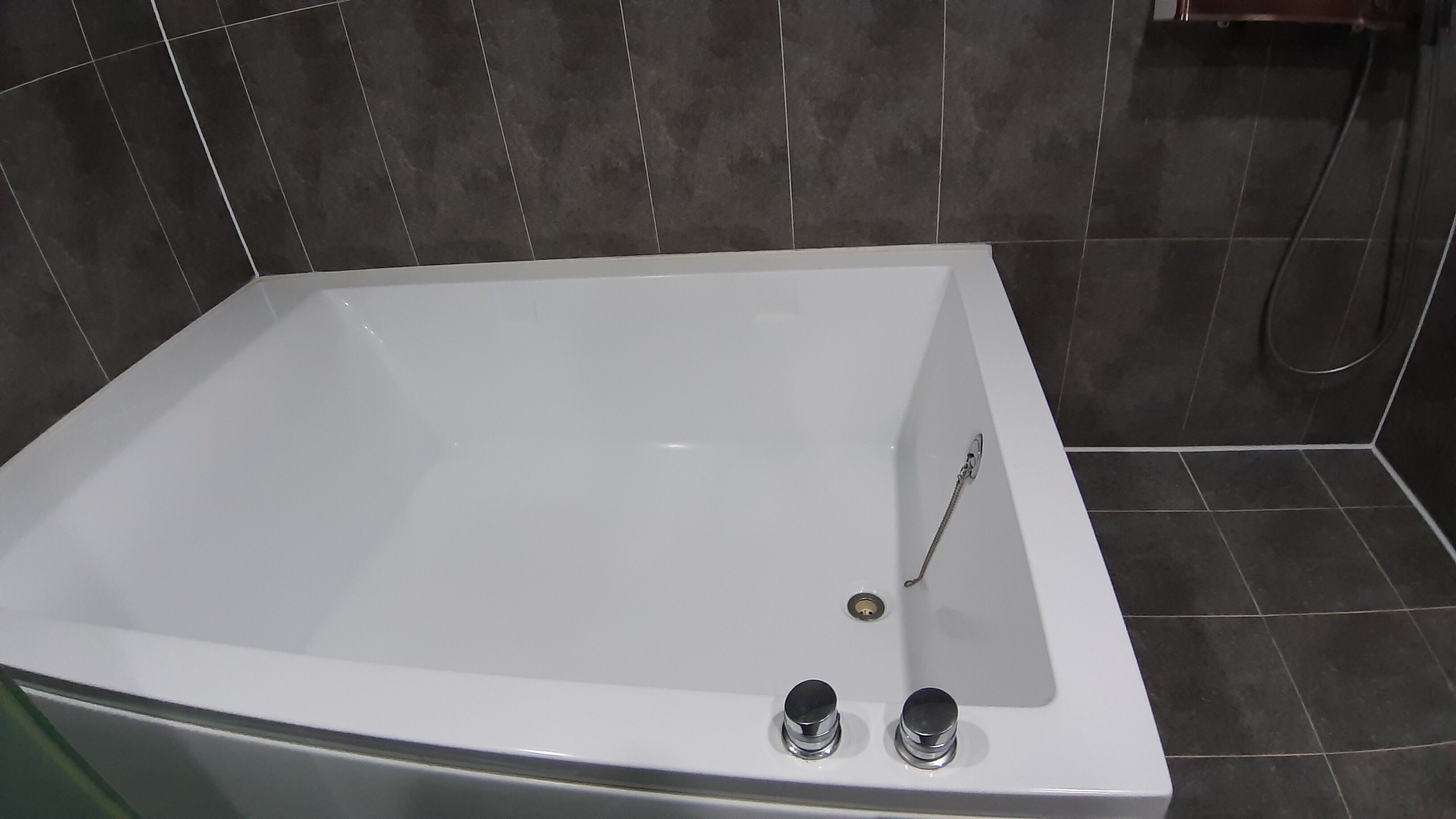 A modern bathroom with a large white bathtub, a shower, and dark tiled walls.