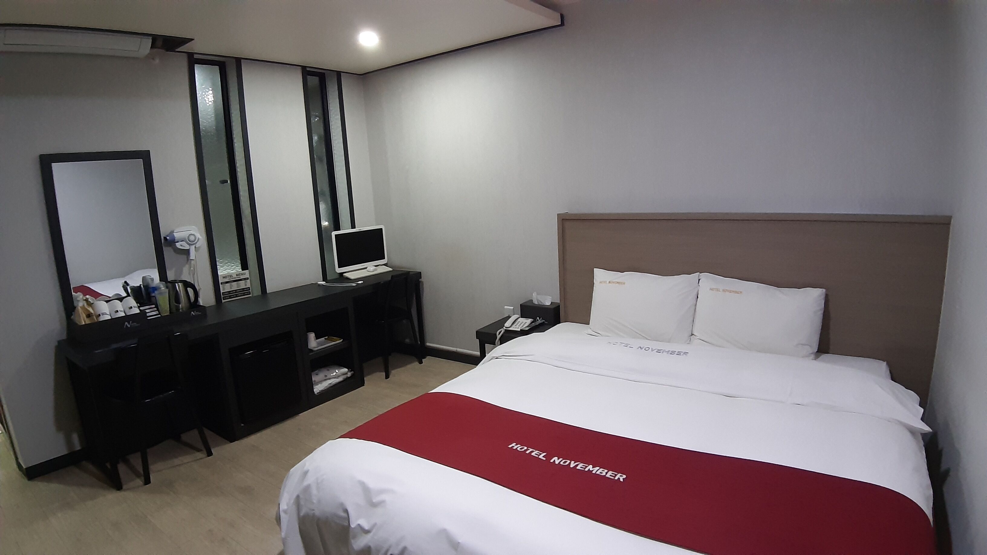 A hotel room with a bed, a desk with a computer, a mirror, and a nightstand with a phone.