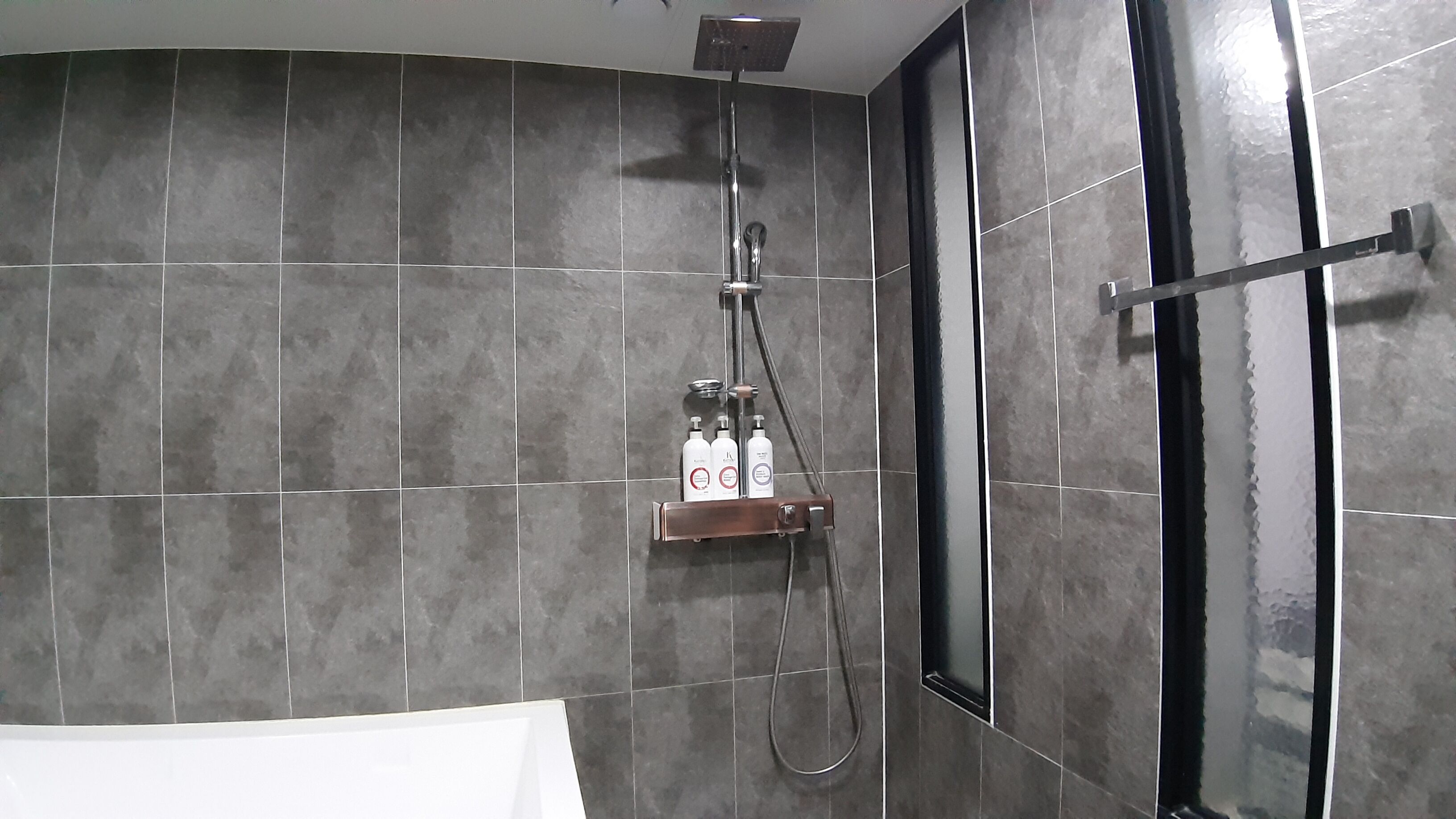 Un baño con paredes de azulejos oscuros, ducha con cabezal de ducha manual y mampara de vidrio.