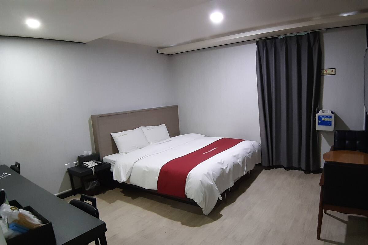 A hotel room with a bed, a desk, a chair, a nightstand, a TV, and a window with curtains.