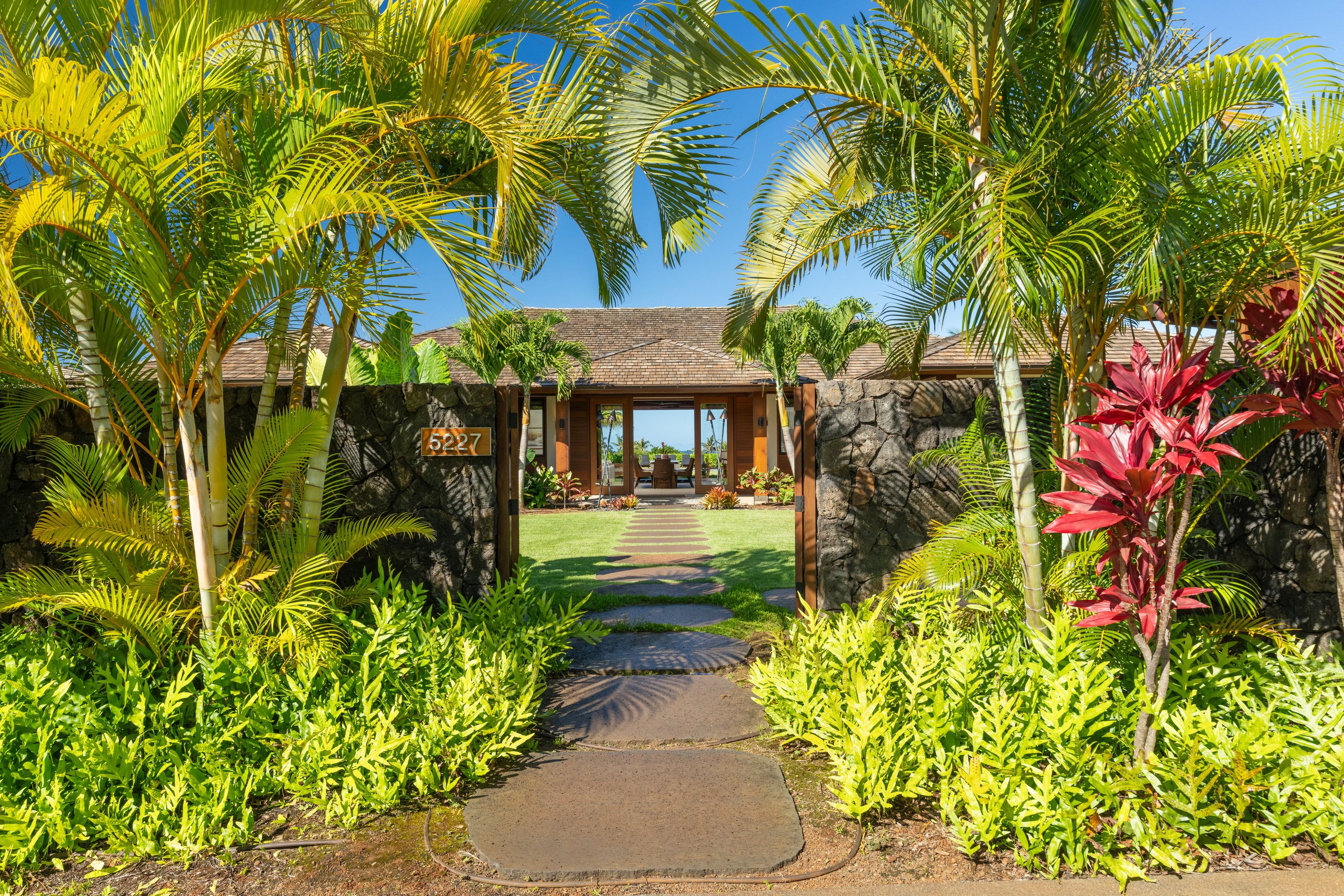 A tropical resort entrance with a stone wall, palm trees, and a paved pathway leading to a building with a tiled roof.