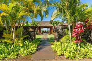 A tropical resort entrance with a stone wall, palm trees, and a paved pathway leading to a building with a tiled roof. - Hale Kipu by Alekona Kauai: Luxury 5br Ocean View Home w Pool, Fire pit, Spa (Koloa)