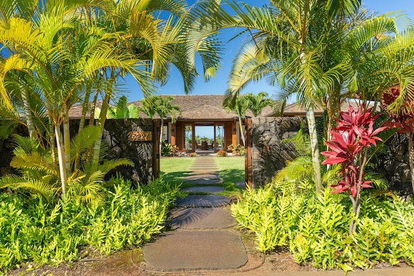 A tropical resort entrance with a stone wall, palm trees, and a paved pathway leading to a building with a tiled roof.