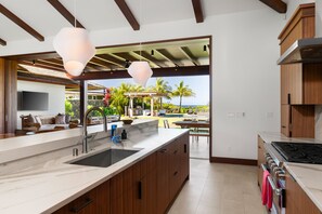 A modern kitchen with a large island, wooden cabinets, and a view of a pool and palm trees.