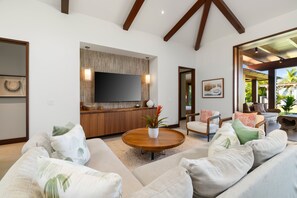 A modern living room with a large flat-screen TV, wooden accents, and a view of the outdoors. - Hale Kipu by Alekona Kauai: Luxury 5br Ocean View Home w Pool, Fire pit, Spa (Koloa)
