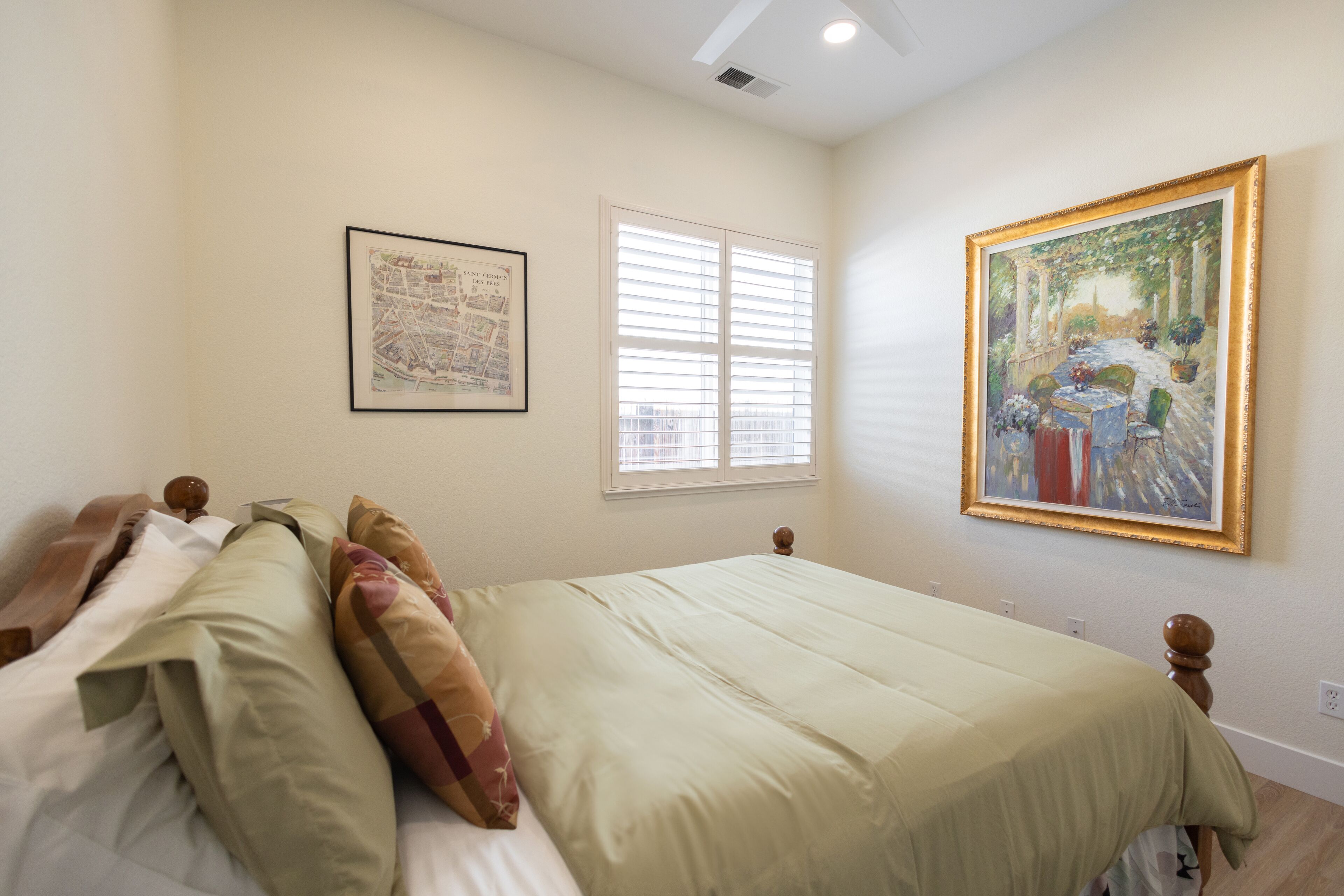A bedroom with a bed, a window with blinds, a framed map, and a framed painting on the wall.