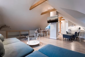 Un loft moderno con techo alto, vigas de madera a la vista, zona de comedor con mesa y sillas, sofá y televisor montado sobre un pilar blanco.