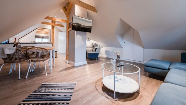 Un loft amplio con techos altos, vigas de madera a la vista, una zona de comedor con sillas de mimbre, una mesa de centro redonda y un sofá azul.
