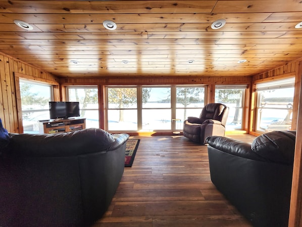 Ein Ferienhausinnenraum mit Holzbalkendecke, einem Fernseher, Ledersofas und großen Fenstern.
