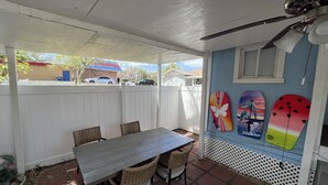 Un'area patio coperta con tavolo e sedie, una recinzione bianca e tavole da surf appese al muro.