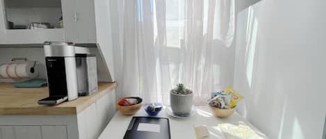 Una habitación pequeña y luminosa con un escritorio blanco, una cafetera, un cuenco, una planta en maceta y una ventana con cortinas traslúcidas.