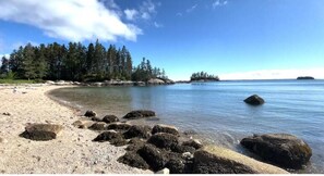 A beach with clear water, large rocks in the foreground, and a forested area in the background. - Cozy Deer Isle Farmhouse Retreat (Deer Isle)