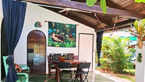 Una zona de pati coberta amb una taula de dinar i cadires, una cadira verda amb funda blava, un mural a la paret i una porta que dona a l'exterior.
