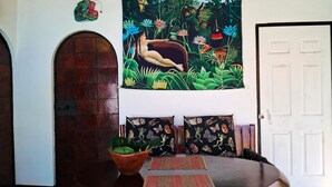 Una habitació amb una taula rodona de fusta, un gran tapís amb una escena de selva i dos coixins estampats.