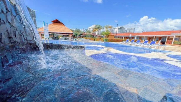 A swimming pool with a waterfall feature, lounge chairs, and a building with a red roof. - Villa Banano 09, Near to the beach with Pool (Jacó)