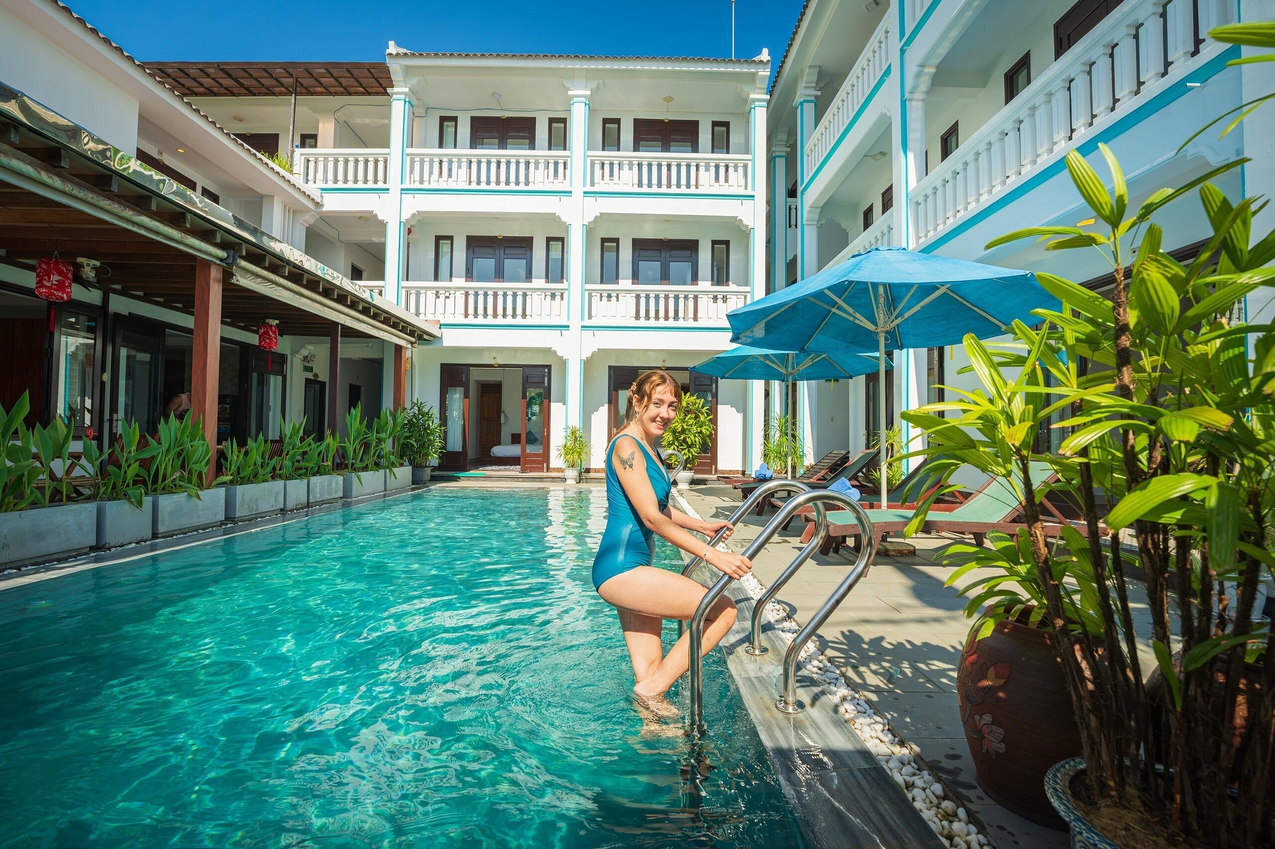 A swimming pool area with a person standing by the poolside, a building with balconies, and potted plants.