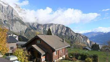 Eine Holzhütte mit geneigtem Dach, umgeben von einer Wiese und Bergen im Hintergrund.