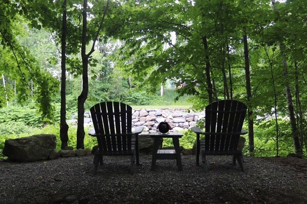 Two Adirondack chairs facing a stone wall with a gravel path and a forested area in the background.