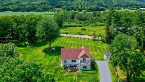 A house with a red roof, surrounded by a well-maintained lawn and trees, set in a rural area with hills in the background. - Riverbend Retreat: A Three-Story Haven in Stowe (Stowe)