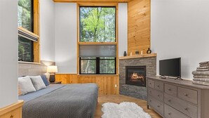 Un dormitorio con cama, chimenea, televisor y un ventanal con vistas a árboles.