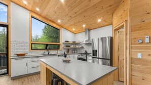 Una cocina moderna con armarios de madera, una isla grande, electrodomésticos de acero inoxidable y ventana con vistas a la vegetación.