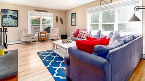 A living room with a grey sofa, red and white pillows, a coffee table, and a blue rug. There are two chairs, a white table, and a window with blinds. - Expansive Stowe home, amazing location and hot tub (Stowe)